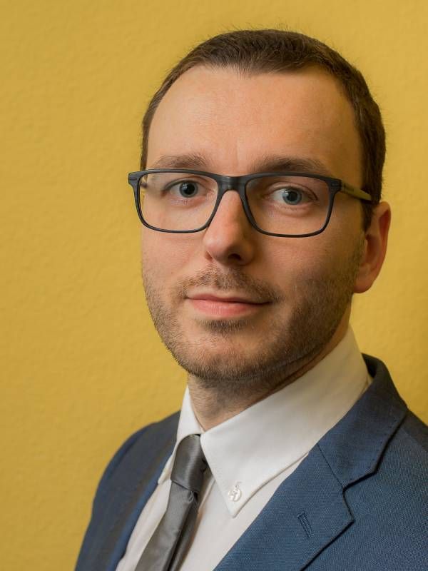 Porträt von Martin Kommer-Hasenest vor gelbem Hintergrund. Er trägt eine Brille und ein blaues Sakko. Unter dem Sakko trägt er ein weißes Hemd, kombiniert mit einer grauen Krawatte.